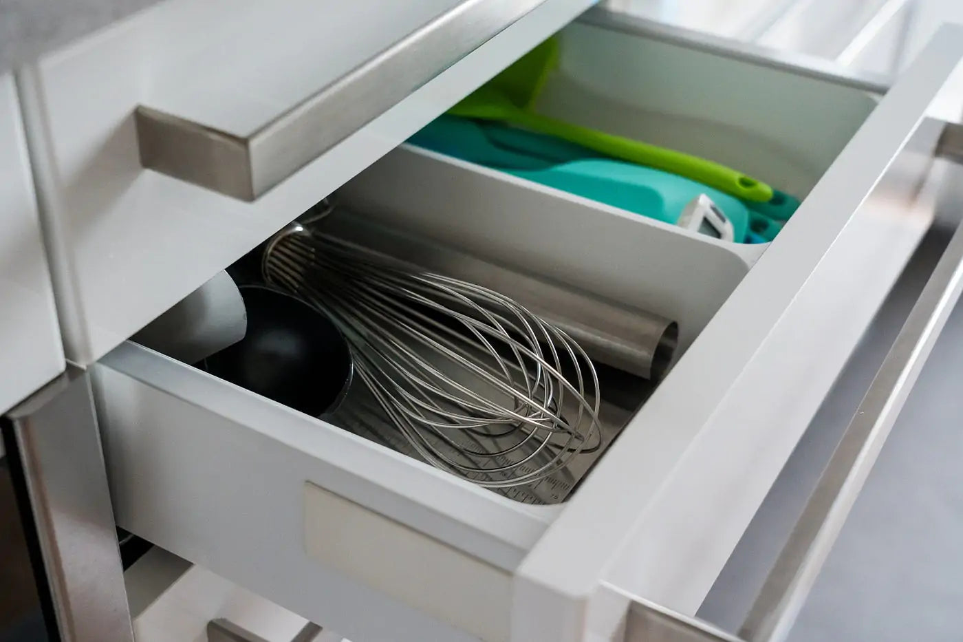 A clean, modern white kitchen drawer equipped with a custom-fit deep utensil divider separating a large metal whisk, measuring cups, and cooking spatulas.
